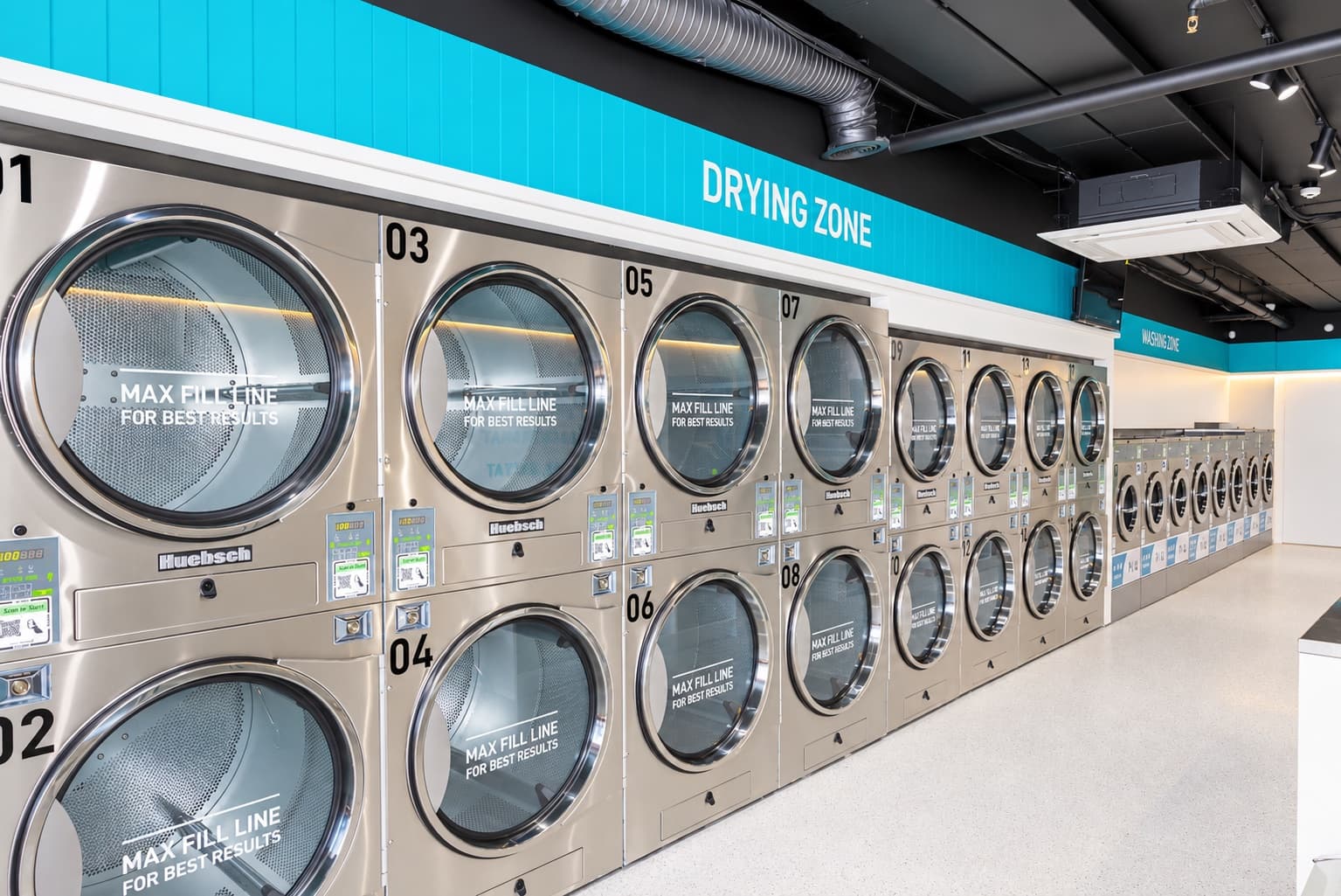 Rows of numbered stacked Huebsch commercial dryers with Max Fill Line door decals beneath the Drying Zone sign, leading back to the washing zone at a Spakleen laundromat