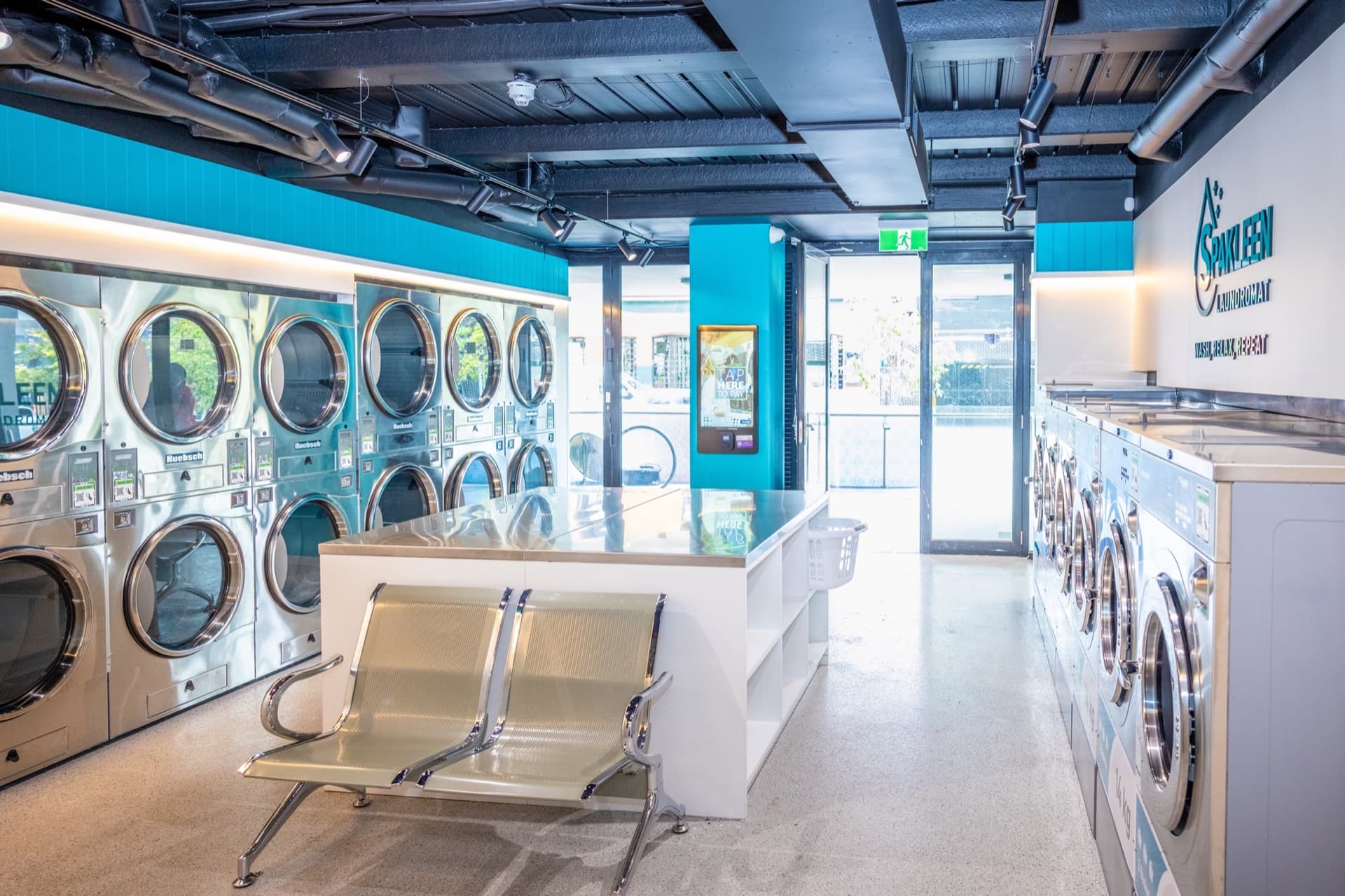 Inside a Spakleen self-service laundromat: stacked commercial dryers, a stainless folding bench with seating, a Tap Here to Pay kiosk by the entrance and a row of washers under the Spakleen Laundromat wall sign