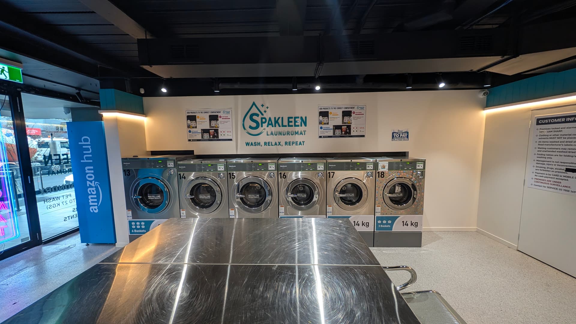 Row of numbered 14kg Spakleen washing machines beneath the Spakleen Laundromat wall sign, with a stainless steel folding bench in the foreground